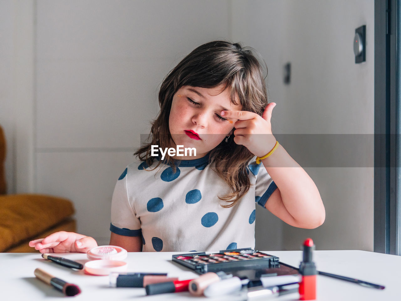 Child with red lips and closed eyes making up eyelid against various beauty products at home