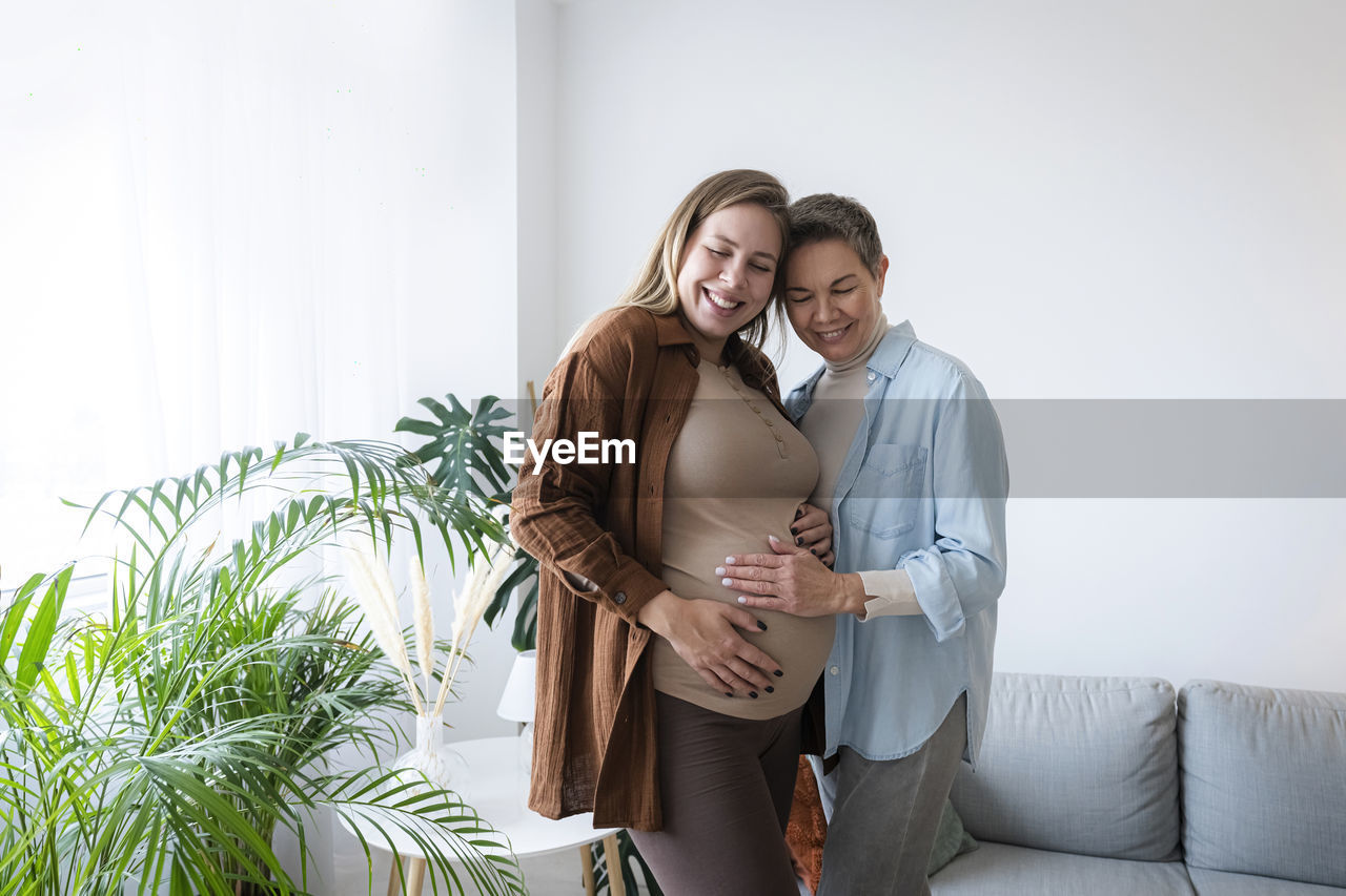 Smiling mother touching belly of pregnant daughter standing at home