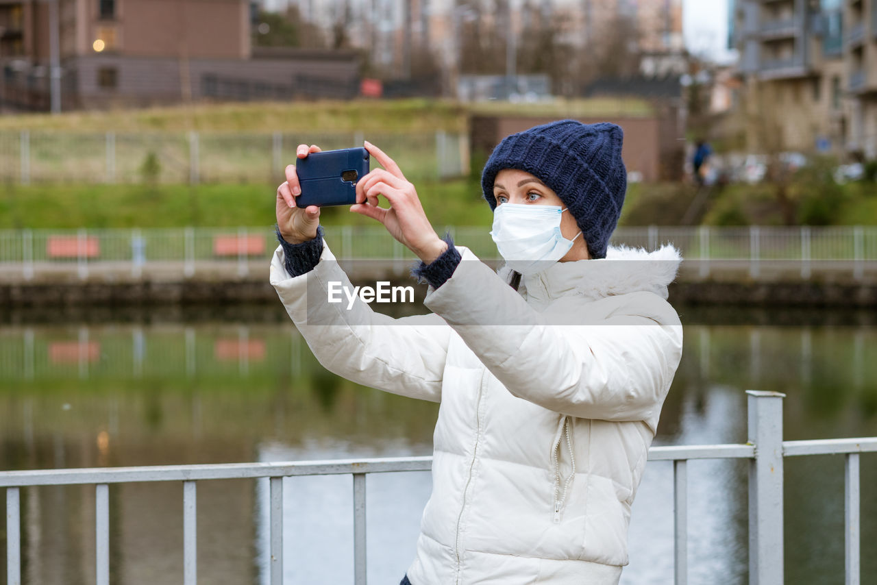 Young woman in a protective mask in a jacket takes pictures on the phone