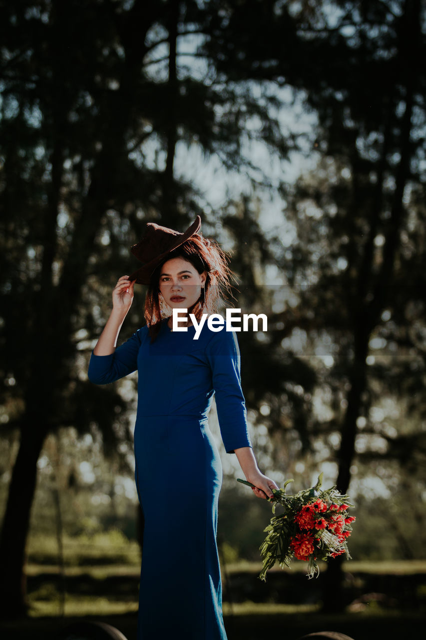 Portrait of young woman holding flower bouquet while standing against trees