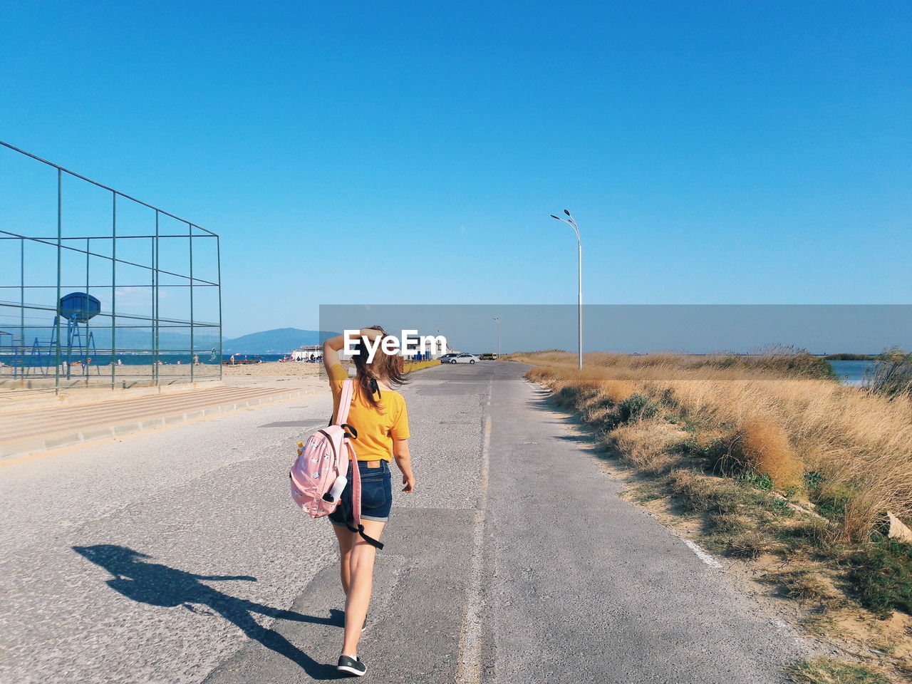 Rear view of backpack woman walking on road against blue sky