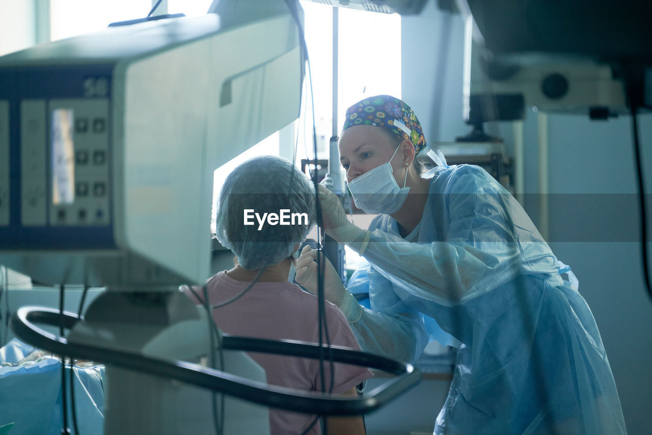 Attentive female surgeon in sterile uniform examining eye of anonymous patient against refractometer in hospital