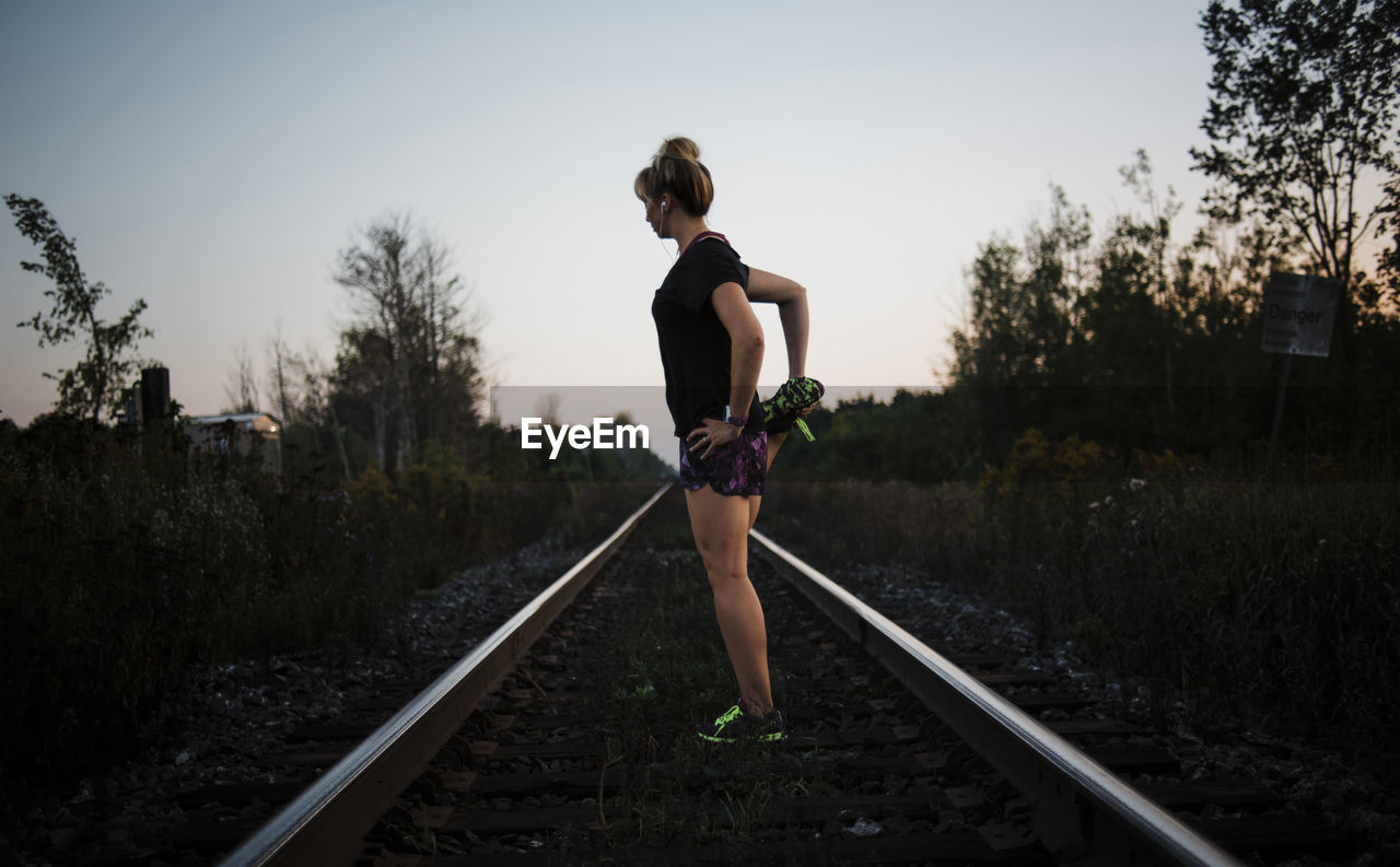 Side view of woman stretching leg while standing on railroad track against sky during dusk