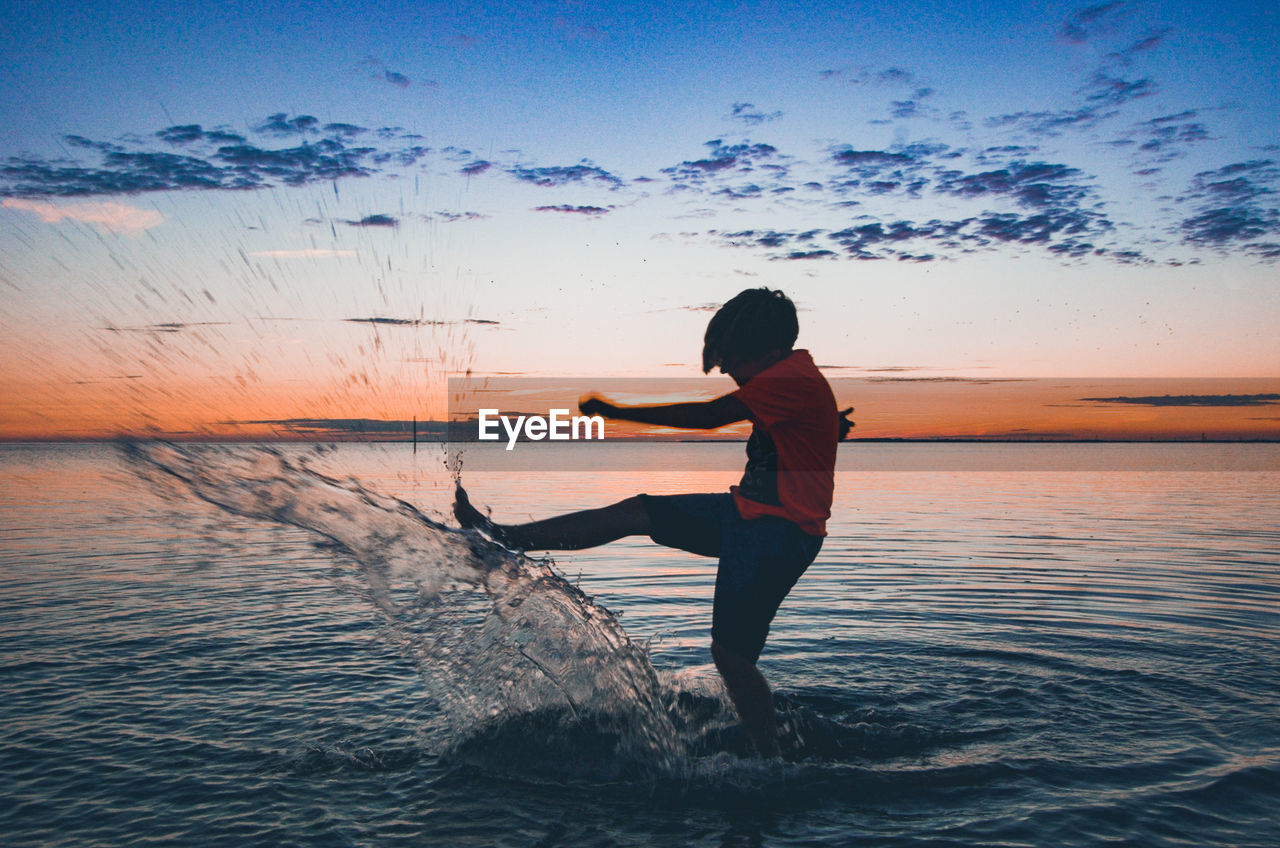 Boy splashing water in sea against sky during sunset