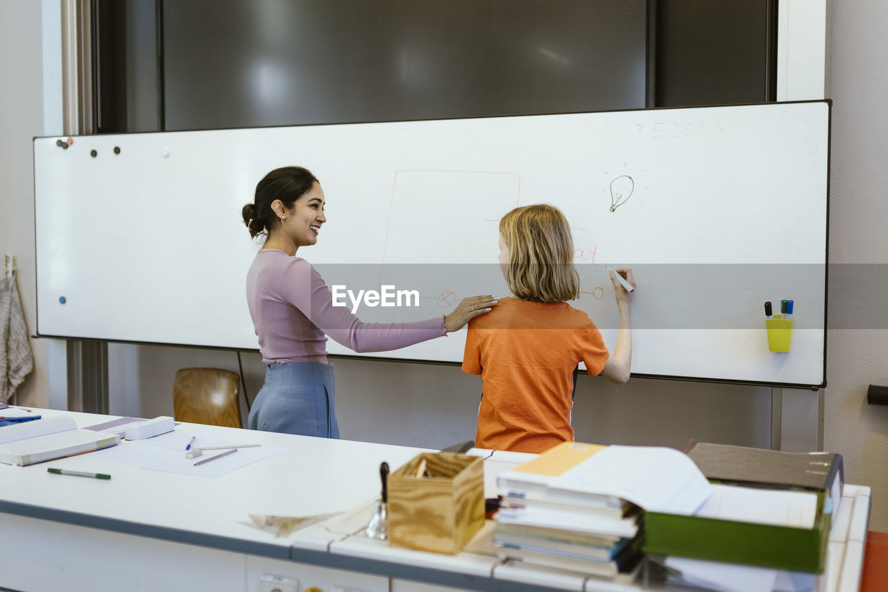 Smiling female student with hand on shoulder of student writing on whiteboard in classroom