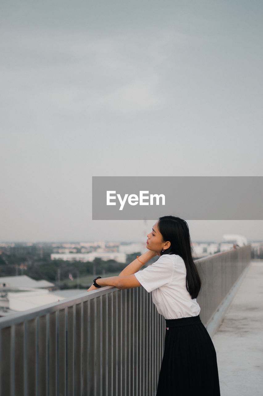 Young woman standing by railing against sky