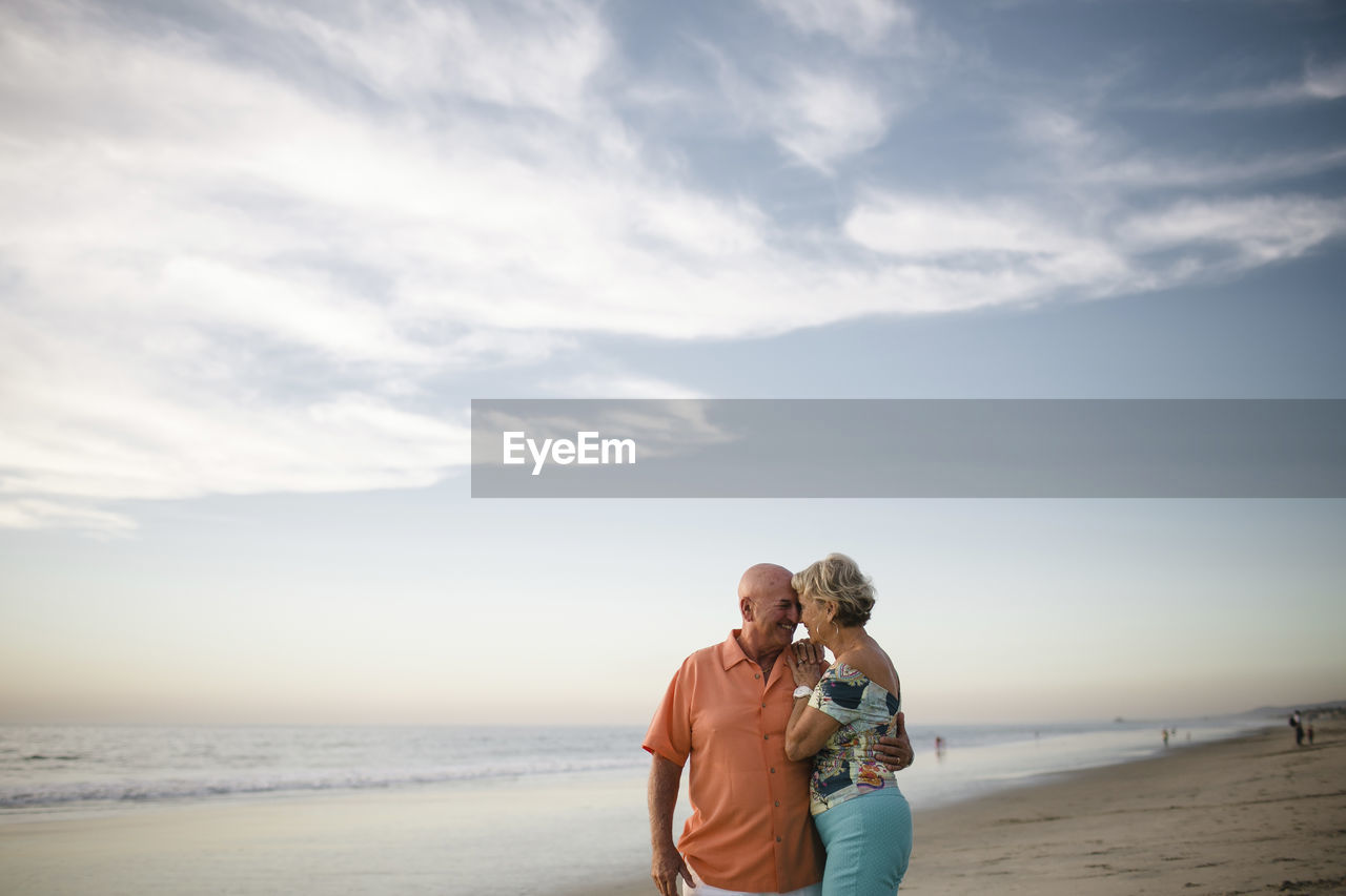 Romantic senior couple looking at each other while standing by sea against sky during sunset