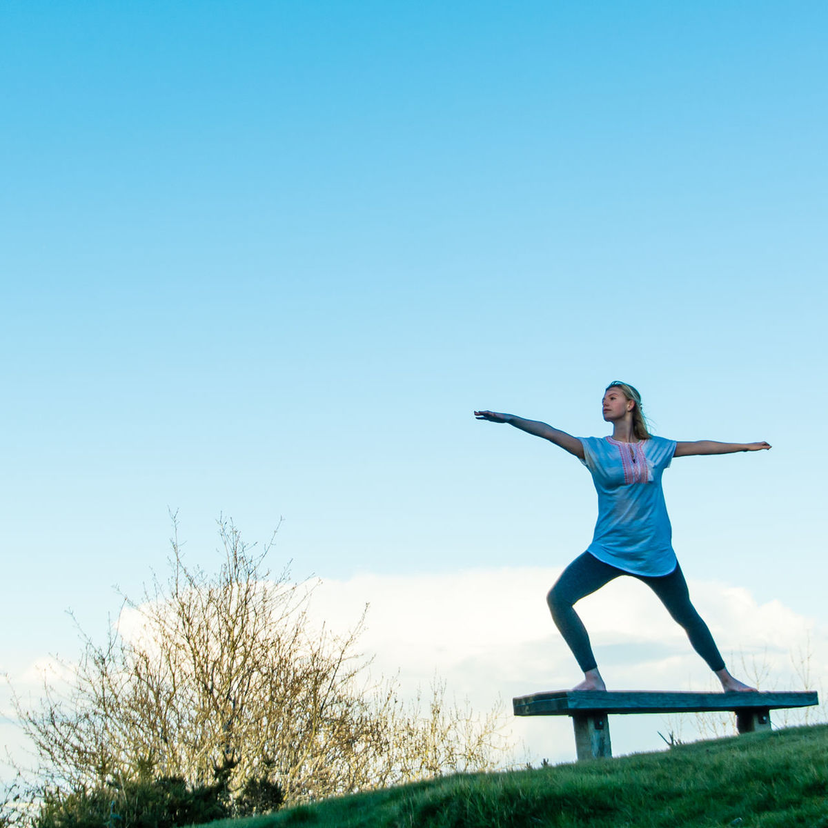 Full length of woman doing yoga on bench at park against blue sky