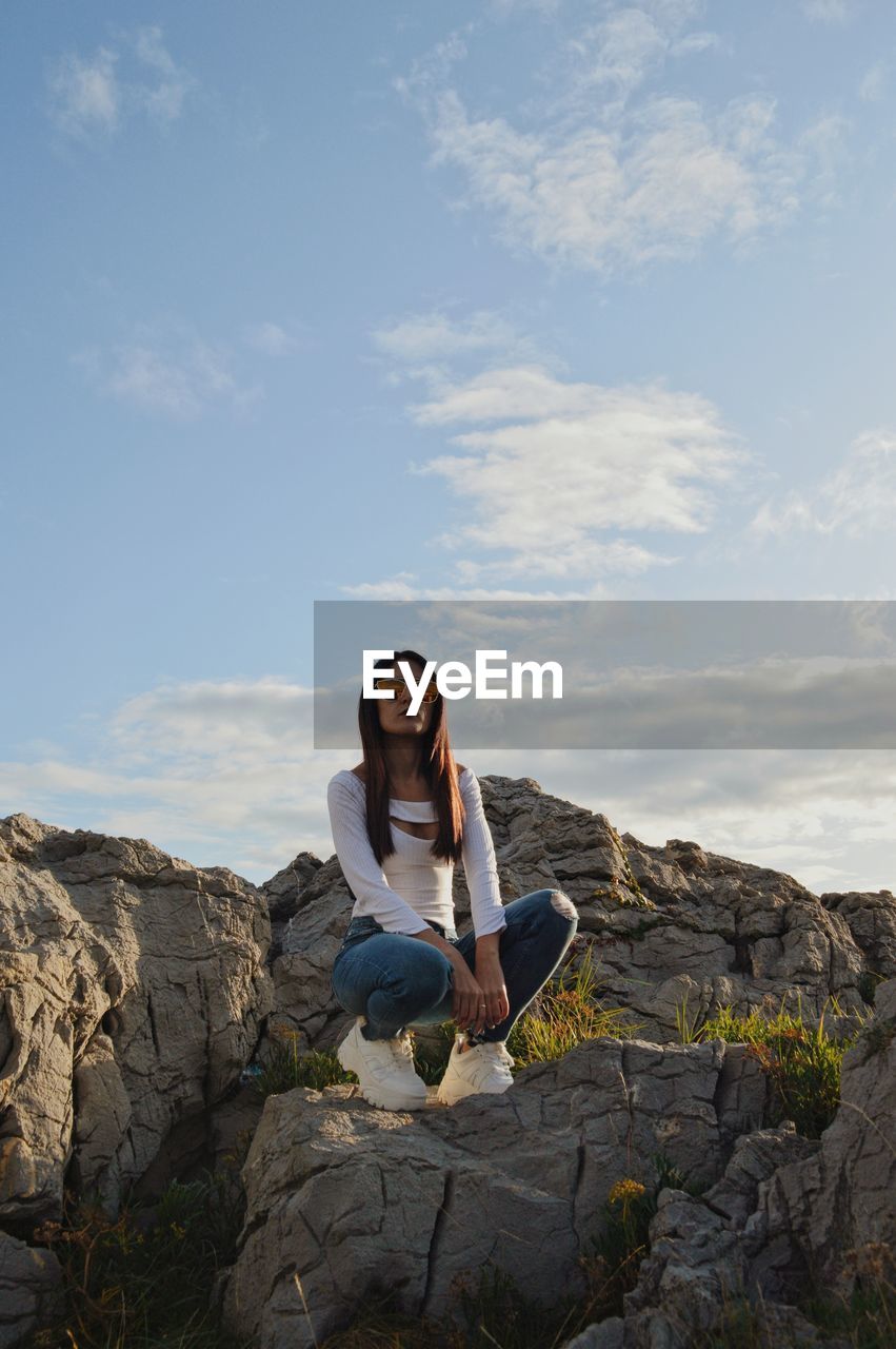 Full length of young woman crouching on rock against sky