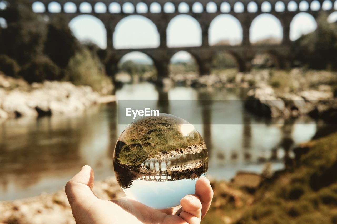 Cropped image of person holding crystal ball with reflection of bridge against sky