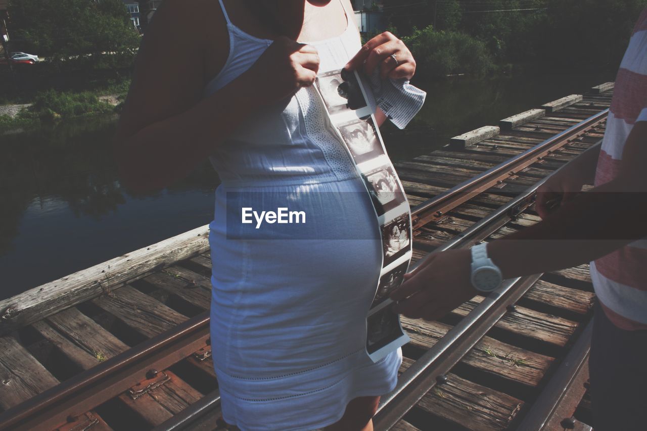 Midsection of pregnant woman with man holding instant print transfer while standing on railway track