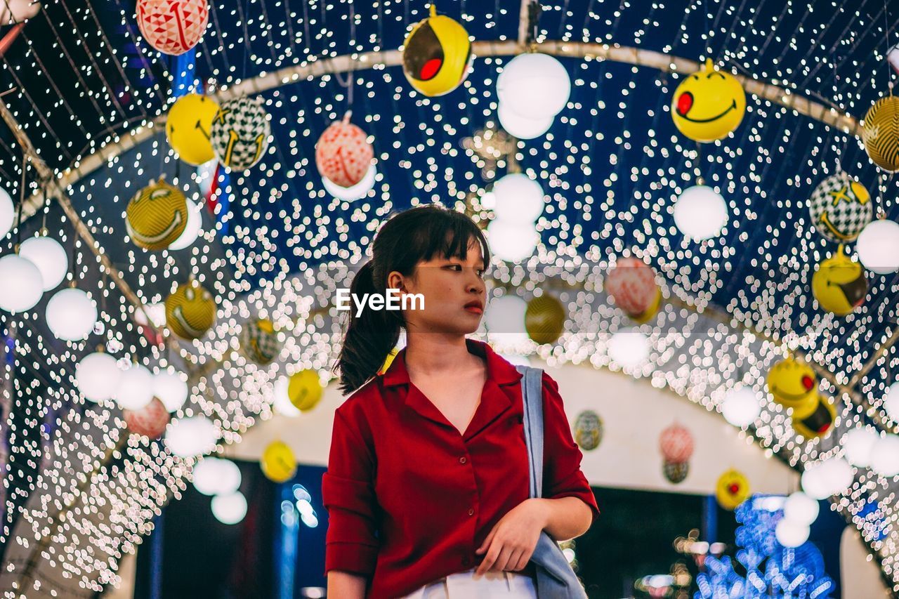 Woman looking away while standing against illuminated decoration at night