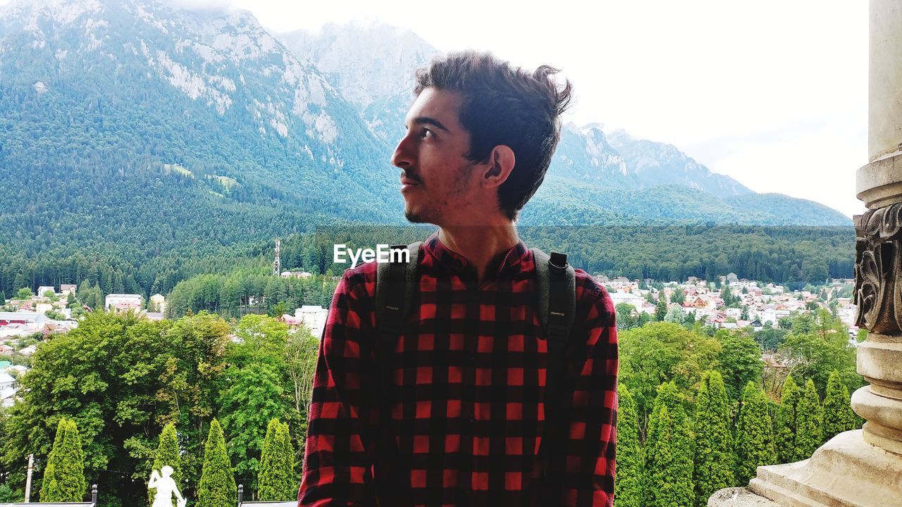 Young man looking away while standing against trees and mountains