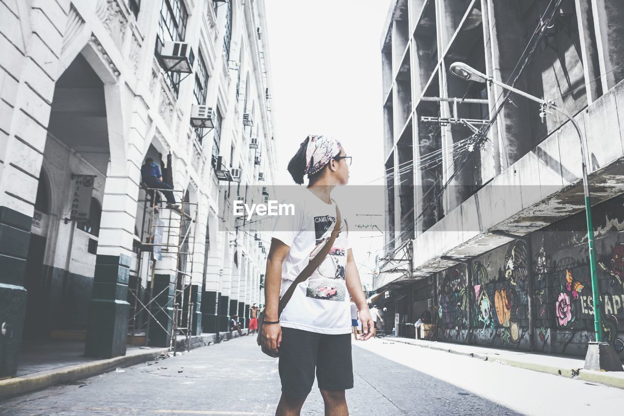 Teenage boy looking away while standing on road amidst buildings in city