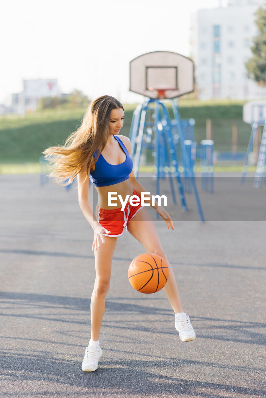 An athletic young woman in sports shorts and t-shirts plays on a basketball court with a ball person