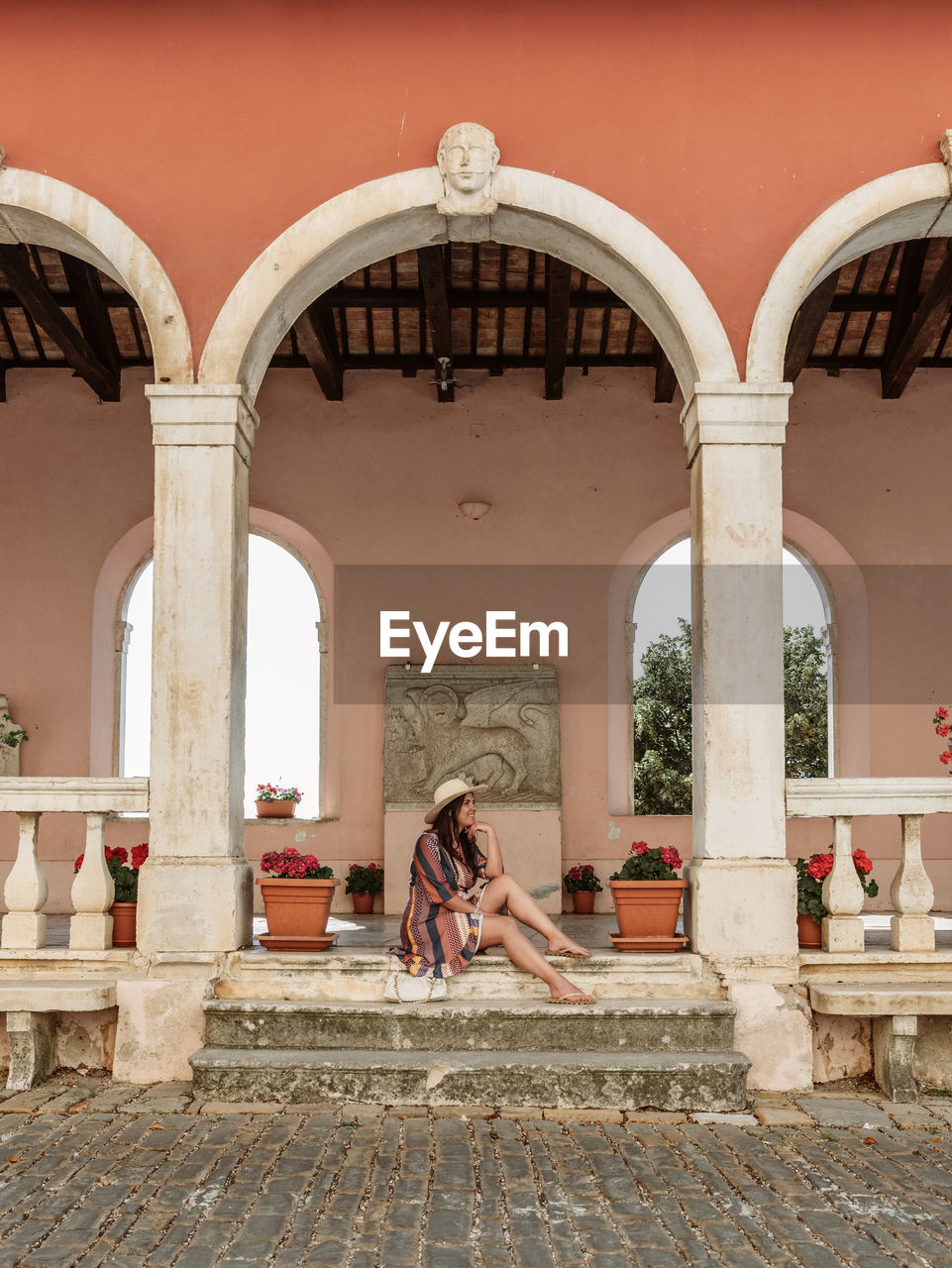 Lifestyle photo of stylish young woman sitting on stone stairs under arched columns of old building