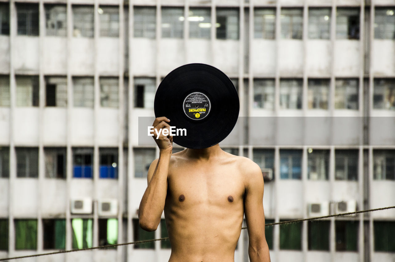 Shirtless man covering his face with record against building