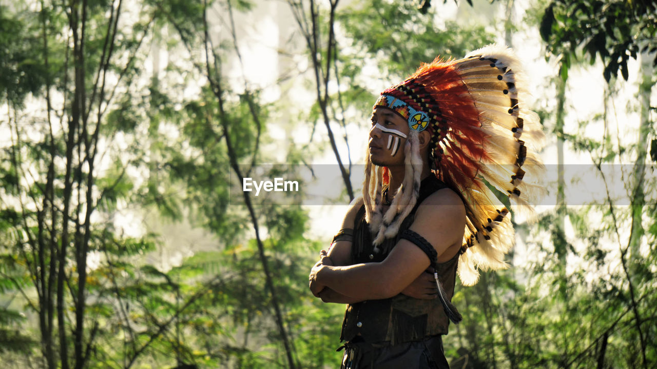 Man wearing feather headdress while standing against trees in forest