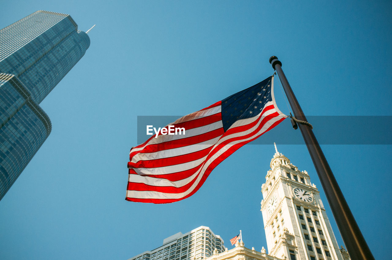 Low angle view of american flag and city buildings against clear blue sky