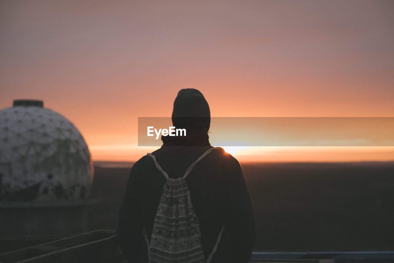 Rear view of silhouette young man with backpack standing against sky during sunset