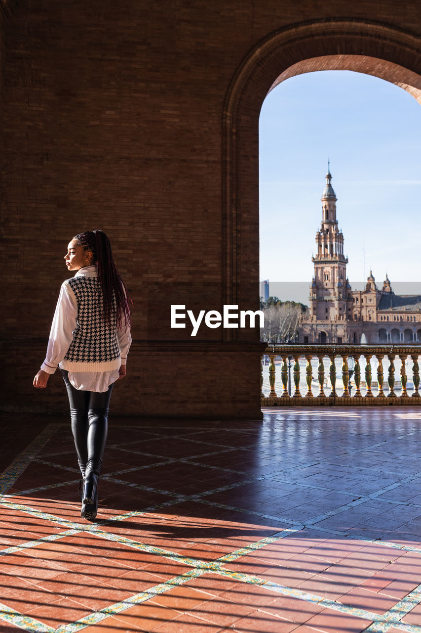 Back view of african american female with braids walking on plaza de espana on sunny day and looking away during weekend in seville