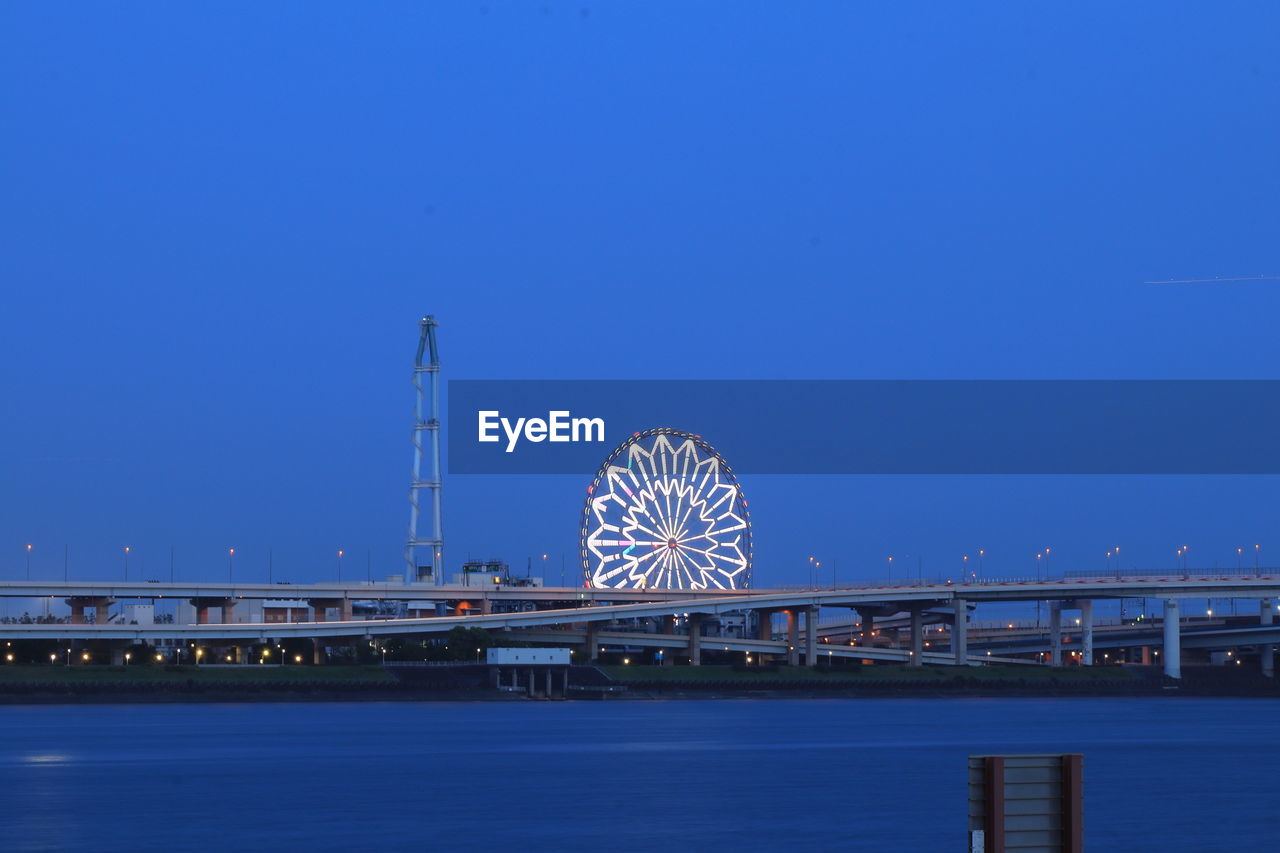 ILLUMINATED FERRIS WHEEL BY RIVER AGAINST CLEAR SKY