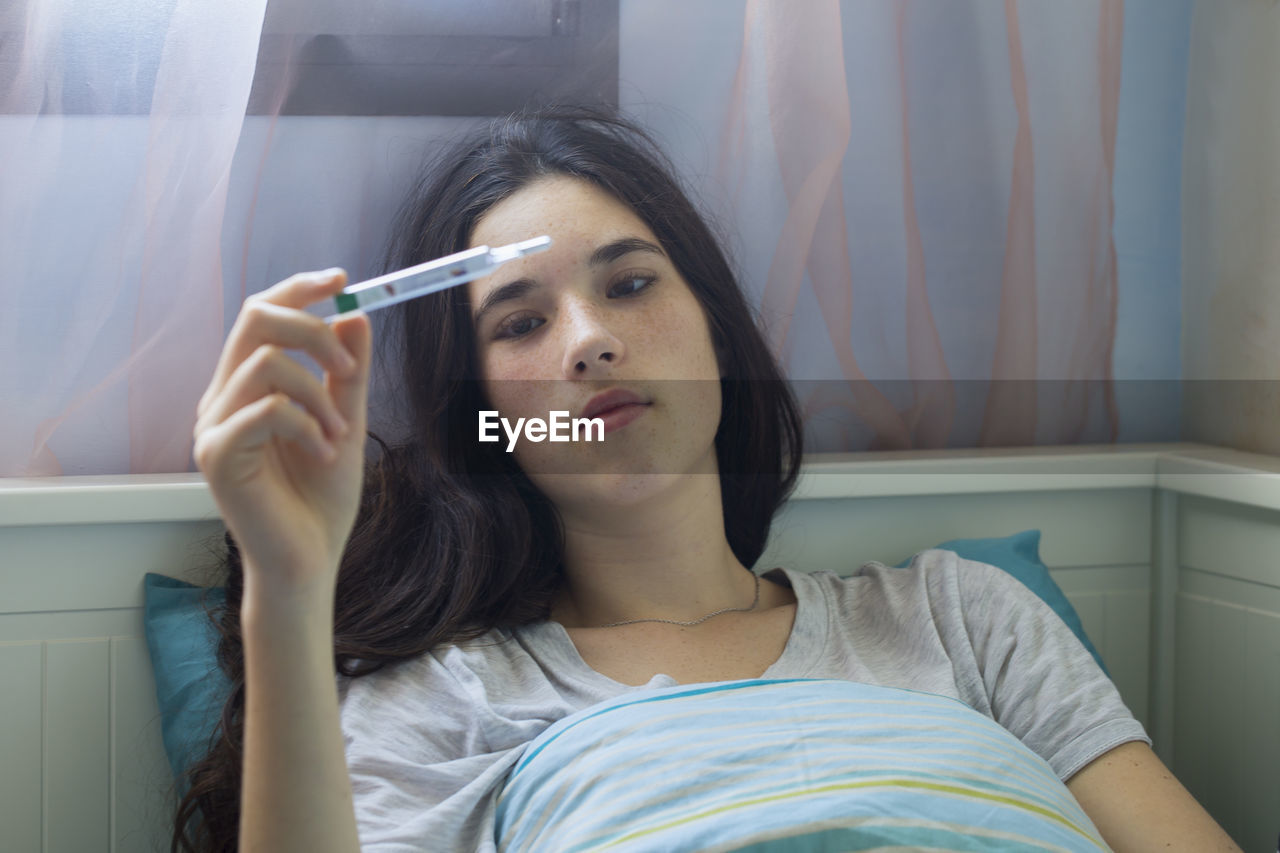 Close-up of teenage girl holding thermometer while lying on bed at home