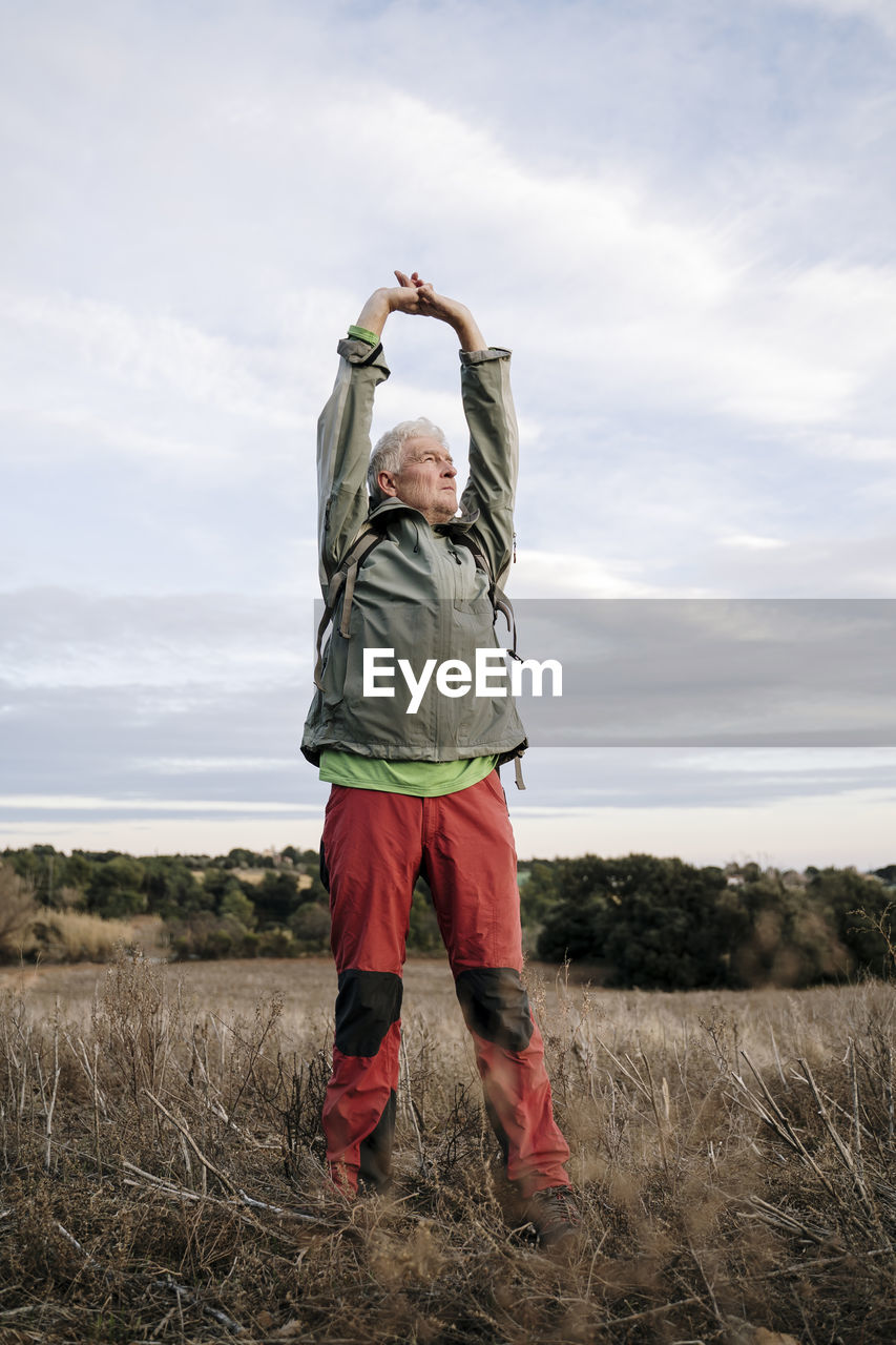 Senior male hiker stretching arms while standing on agricultural field against cloudy sky at countryside