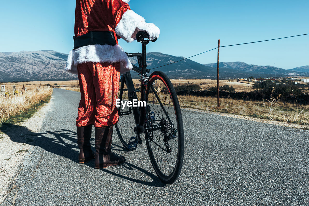 Cropped unrecognizable santa claus in red clothes standing with modern bicycle on empty road and looking away