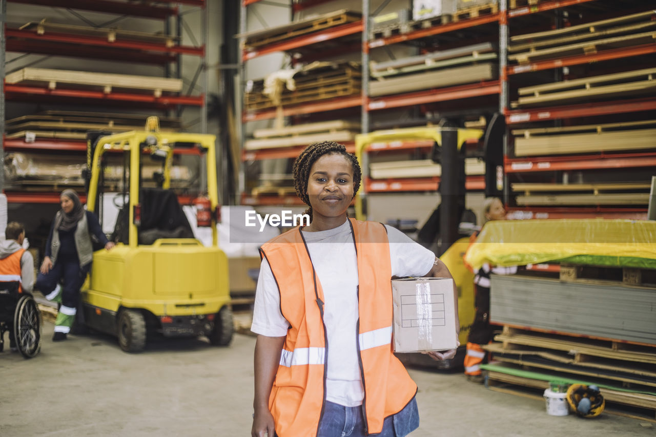 Portrait of smiling carpenter carrying cardboard box in warehouse