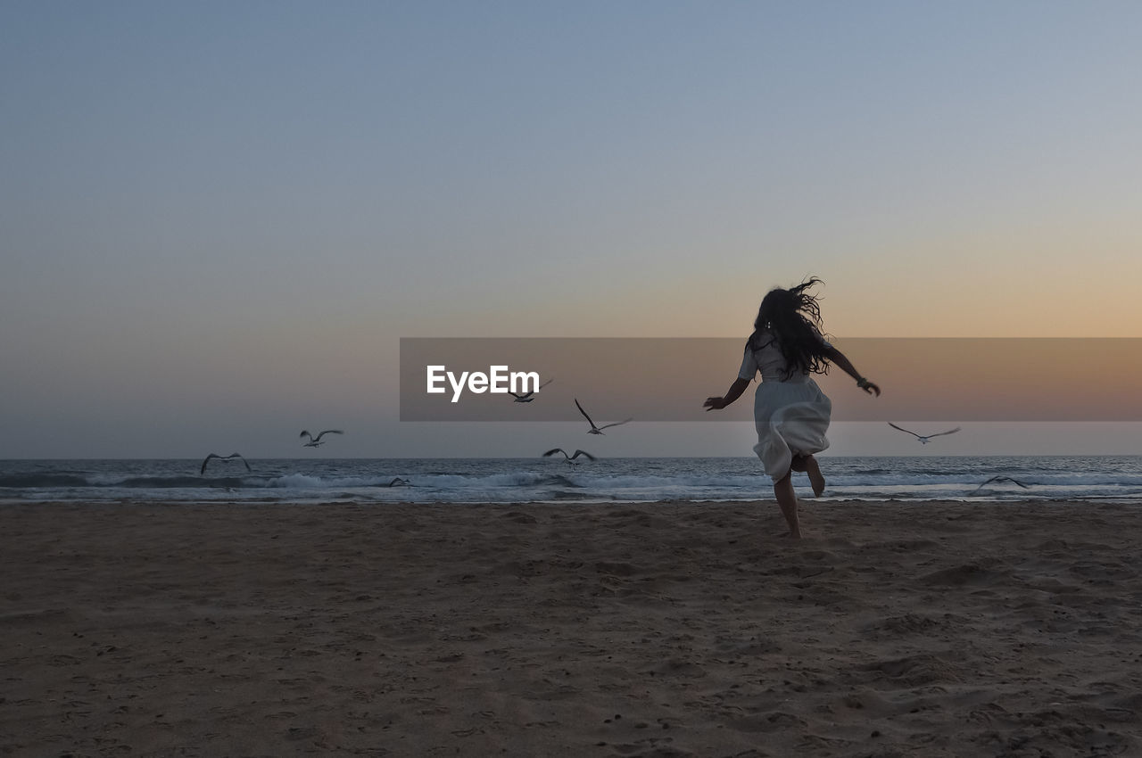 WOMAN ENJOYING ON BEACH AGAINST SKY DURING SUNSET