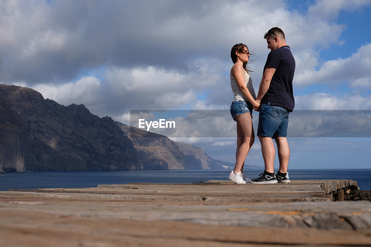 Full length of couple standing on pier over sea against cloudy sky