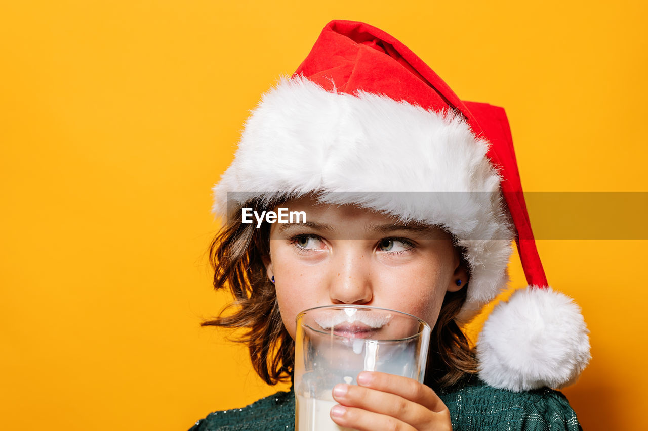 Cute girl in santa hat sipping milk from glass and looking away during christmas celebration against yellow background