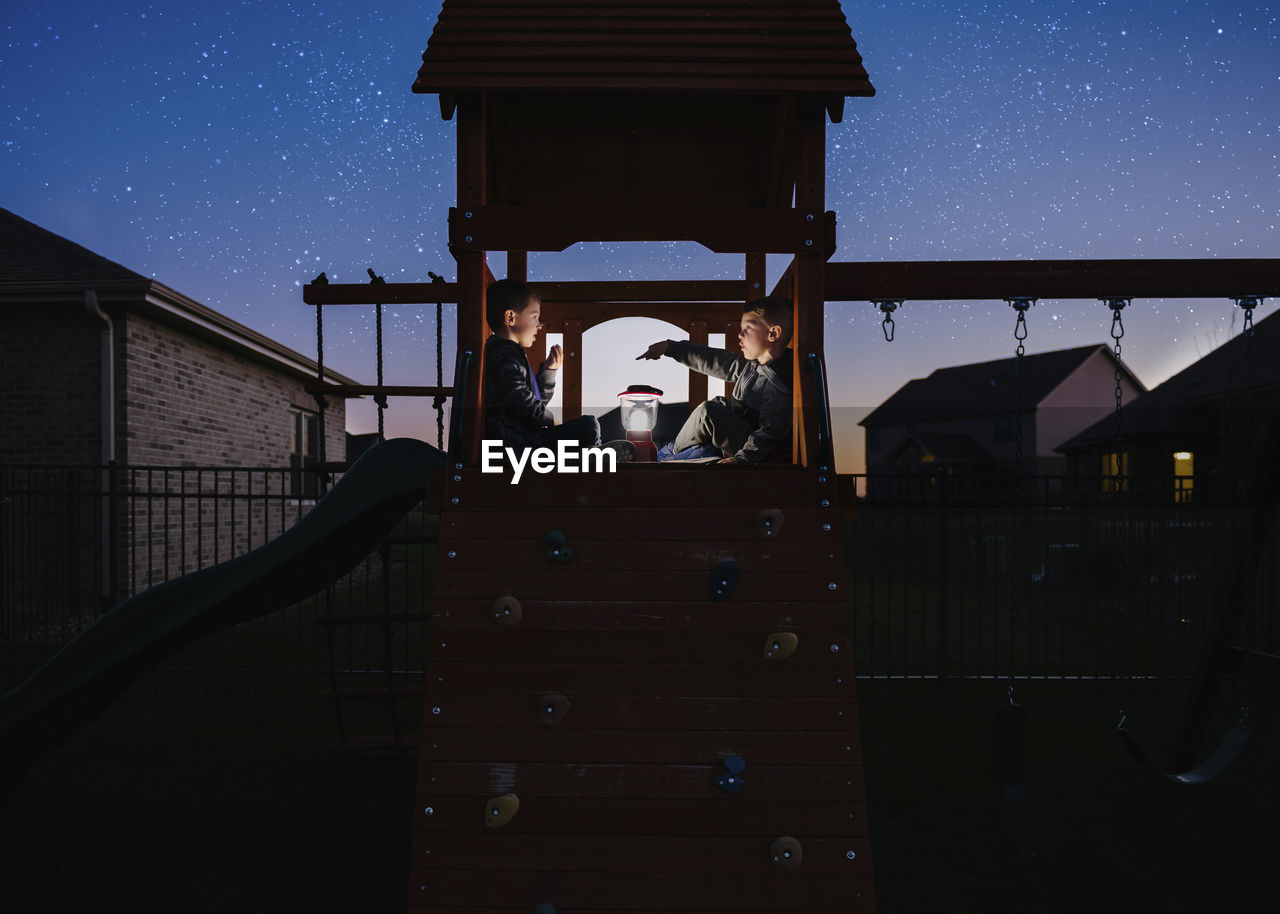 Brothers with illuminated lantern sitting on outdoor play equipment against star field at park