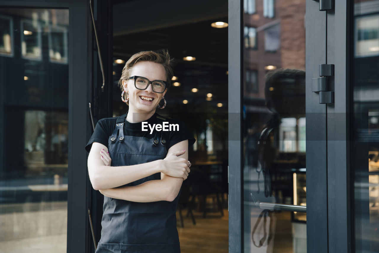Portrait of smiling androgynous owner standing with arms crossed at coffee shop entrance