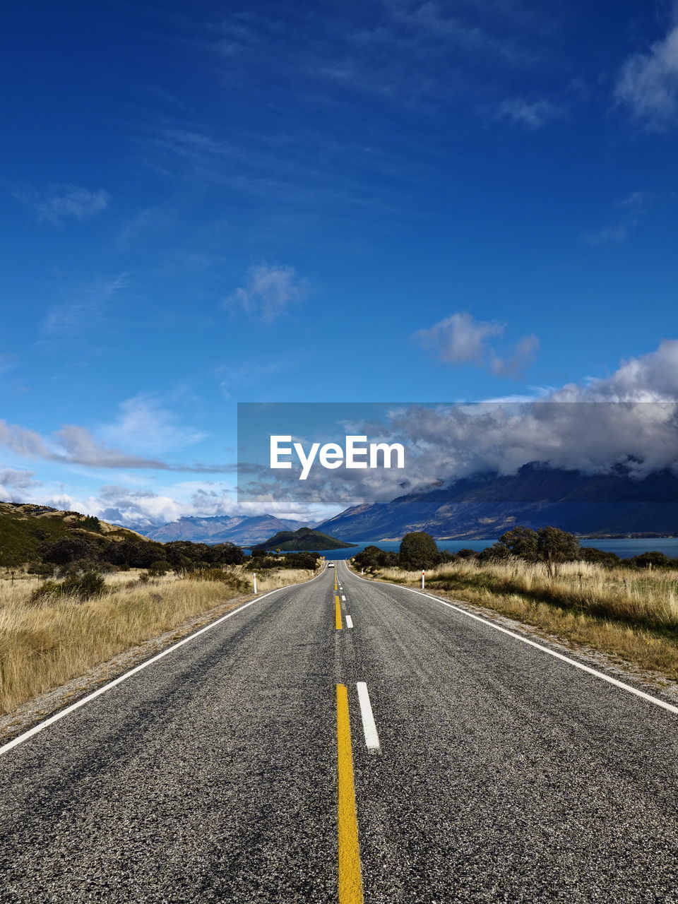 Empty road leading towards lake and mountains against blue sky