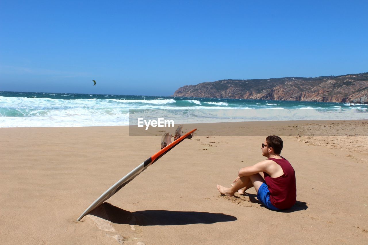 Side view of young man with surfboard sitting at beach against blue sky during sunny day