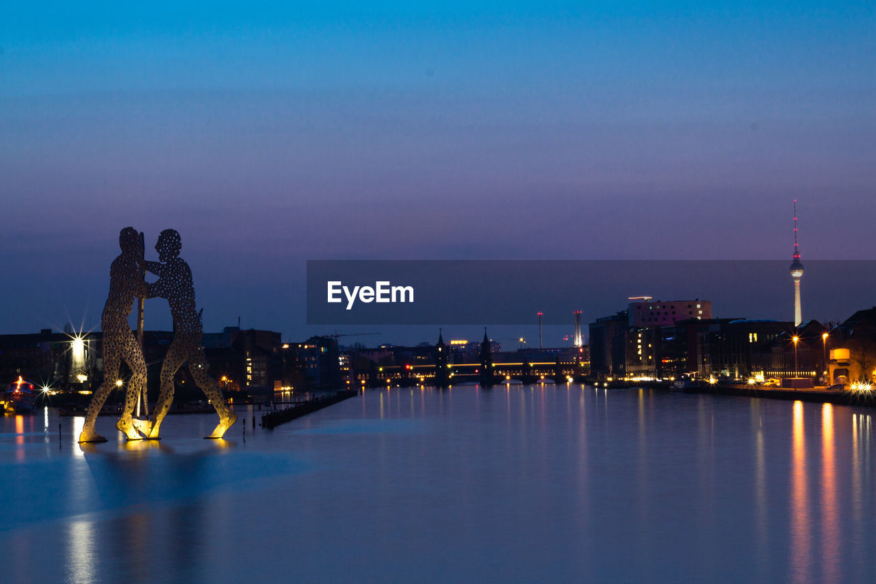 Molecule men sculpture on river in city against sky at night