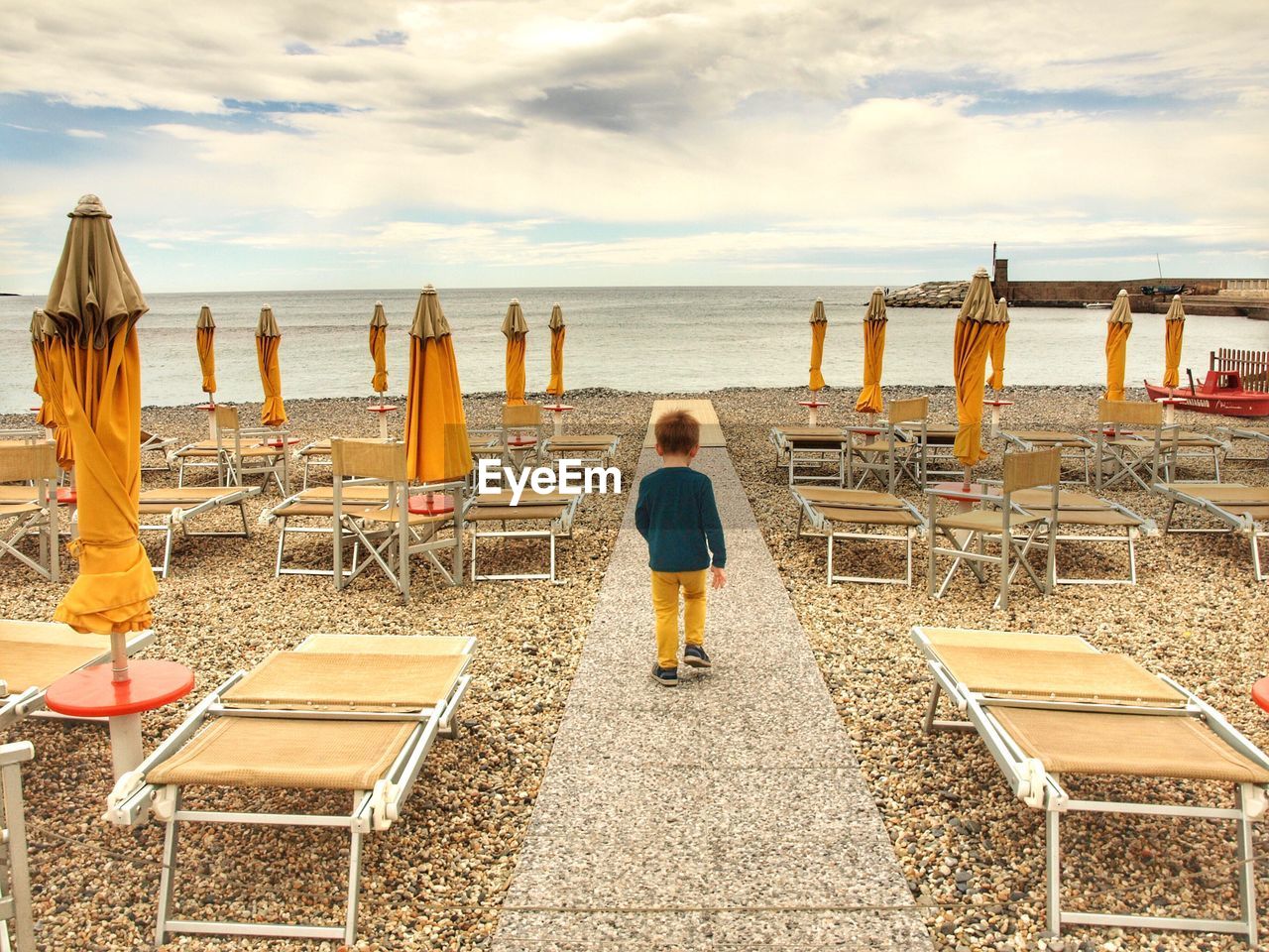 Full length rear view of boy walking on footpath at beach amidst deck chairs and parasols