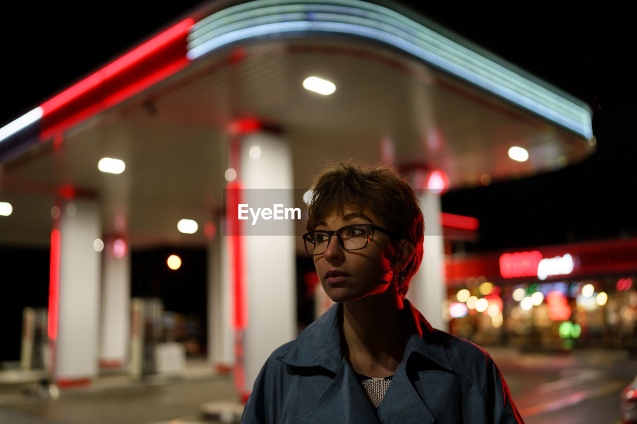 Depressed young woman in glasses stands alone near gas station looks away with sadness at cold night