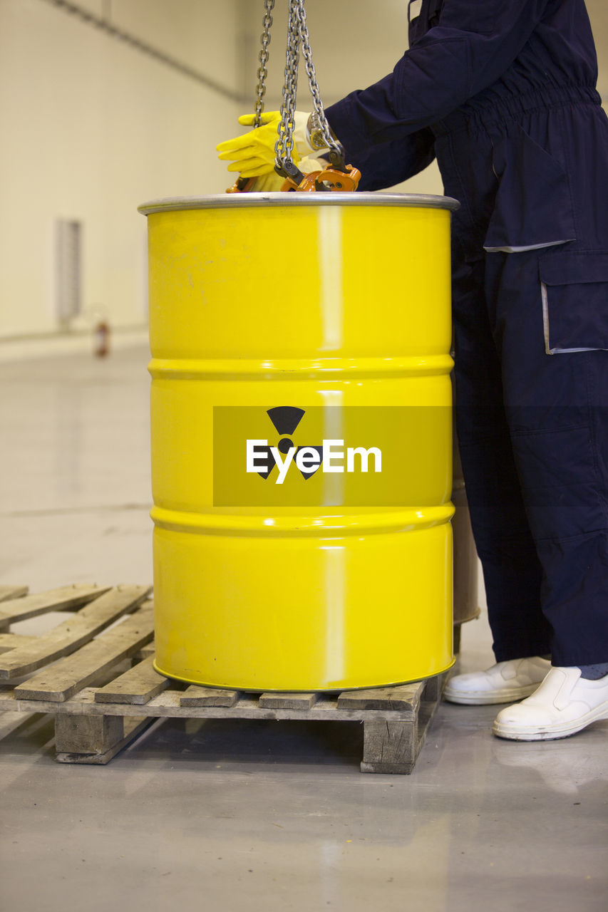 Low section of worker standing by yellow barrel with caution sign in factory