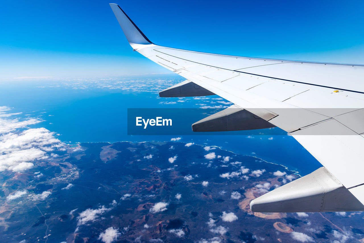 Cropped image of airplane wing flying over sea against blue sky