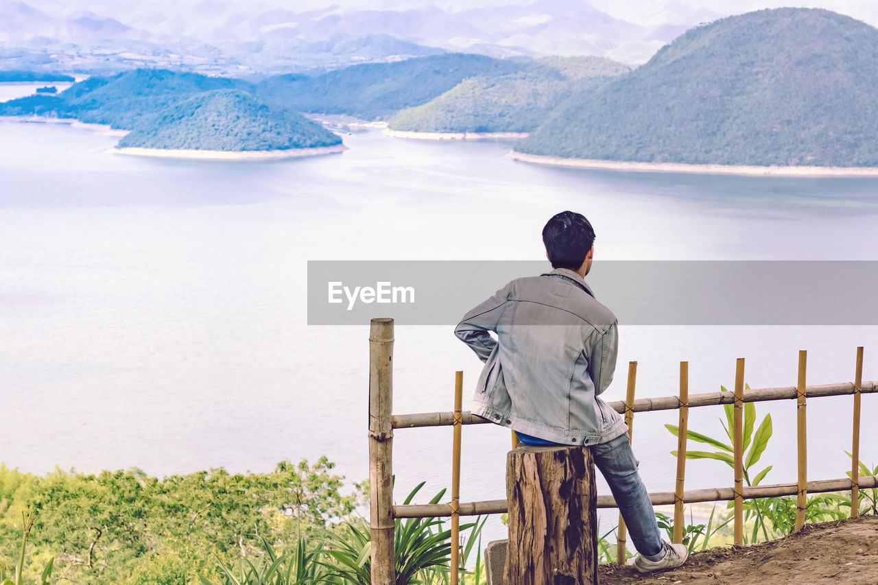 REAR VIEW OF MAN LOOKING AT LAKE BY MOUNTAIN