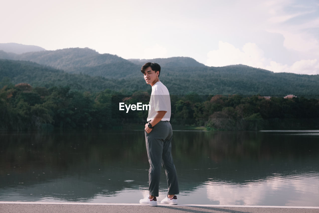 FULL LENGTH OF YOUNG MAN STANDING ON LAKE AGAINST MOUNTAIN