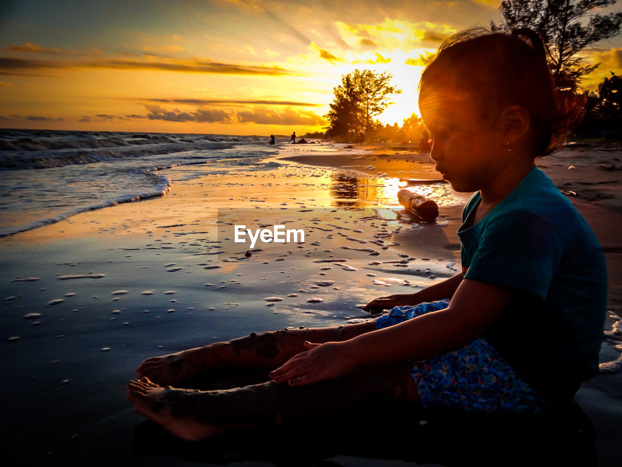 Little girls sitting on shore at beach against sky during sunset