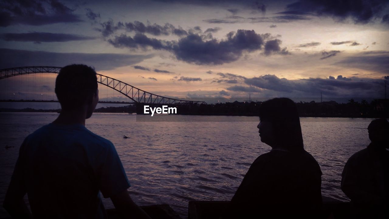 SILHOUETTE PEOPLE ON SUSPENSION BRIDGE OVER SEA AGAINST SKY