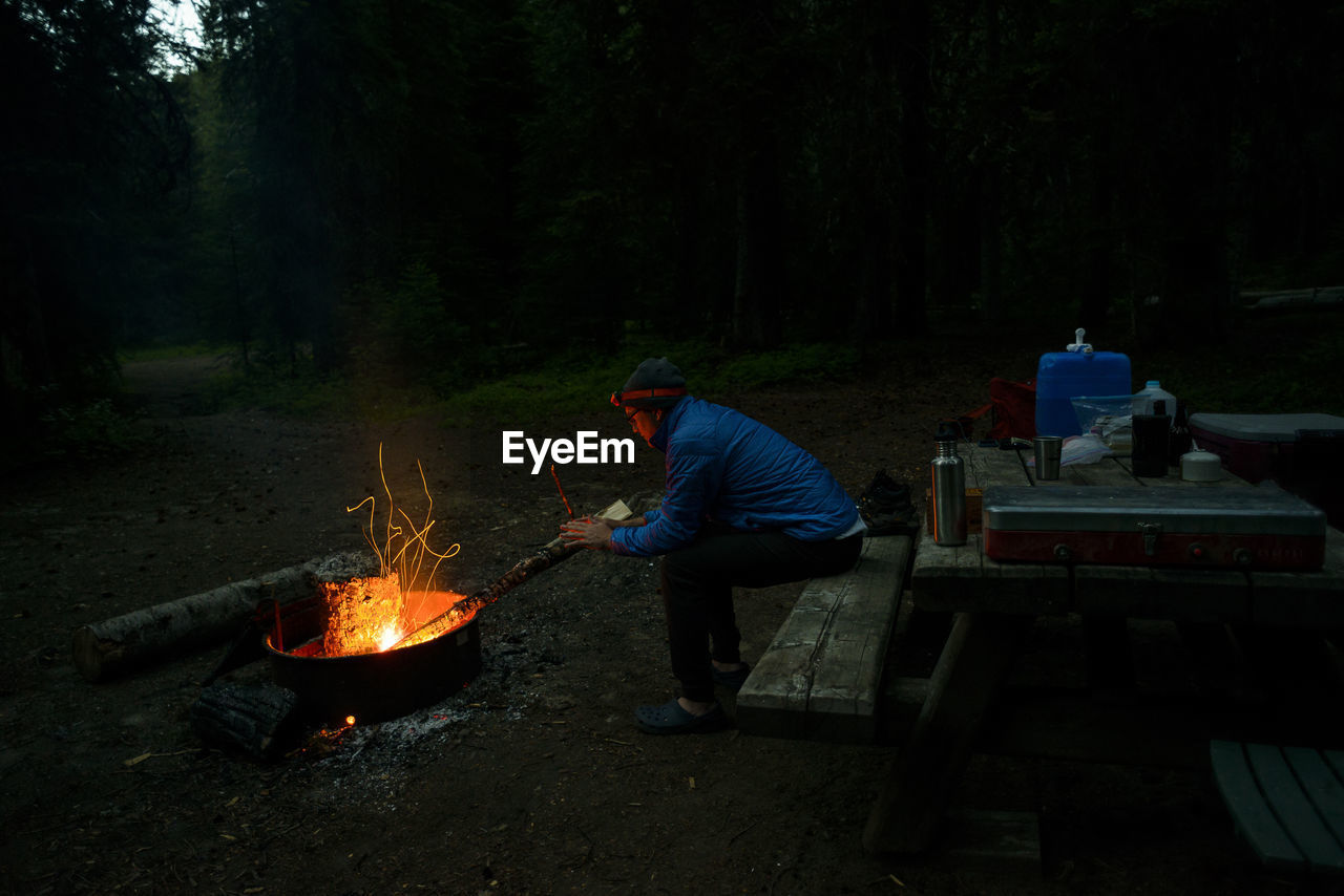 Side view of hiker warming hands by campfire at campsite during dusk