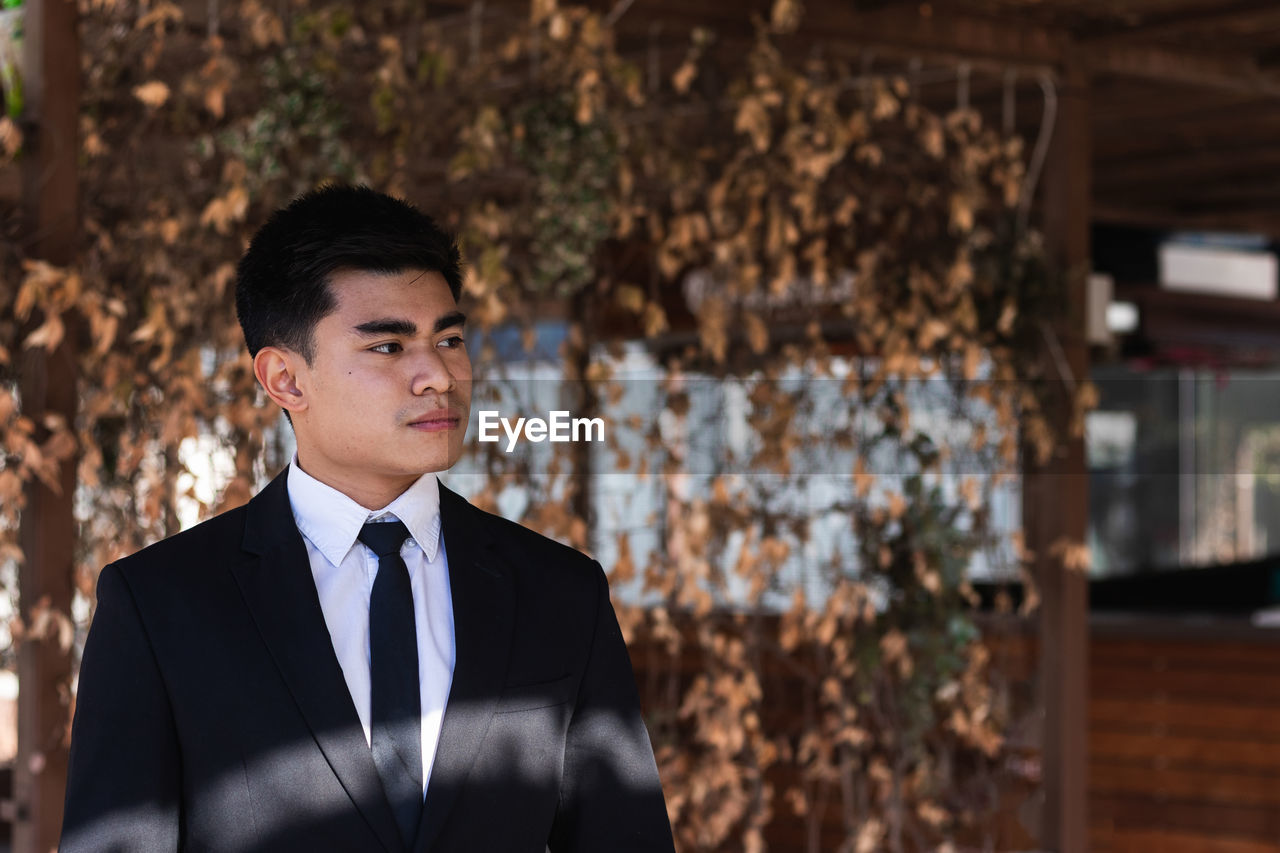 Serious ethnic male entrepreneur wearing classy suit standing in shadow on sunny day in city and looking away