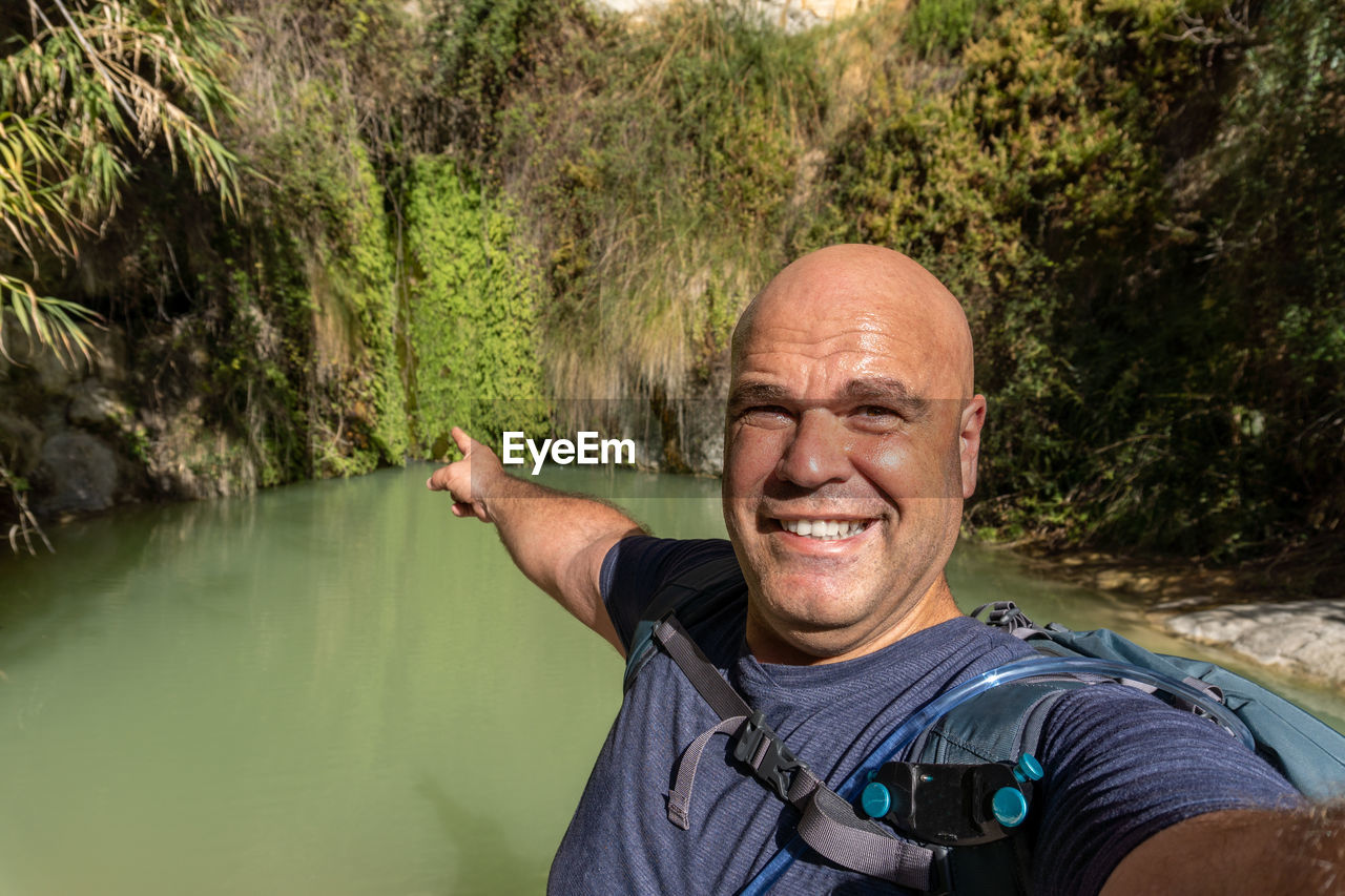 Smiling hiker man takes a selfie pointing to a small natural pool with green water