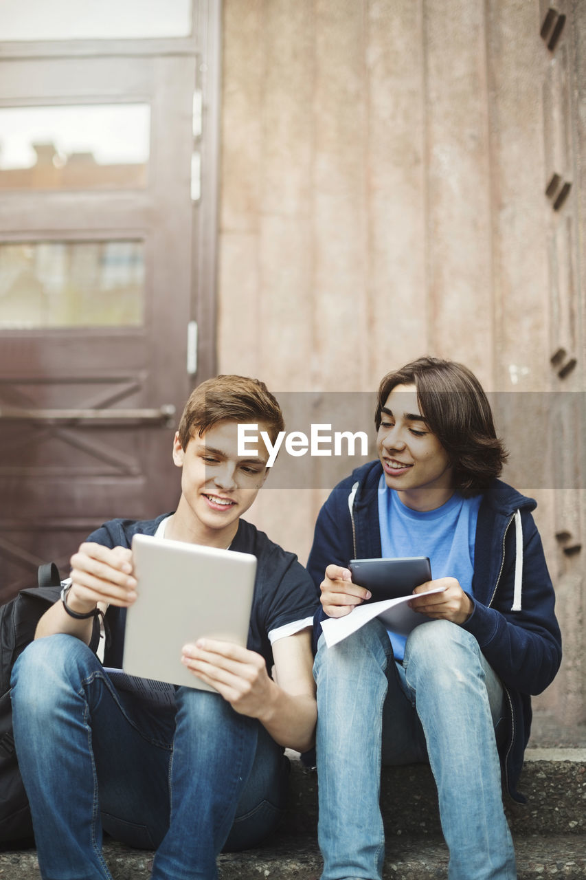 Happy male teenagers using digital tablet while sitting on steps outdoors